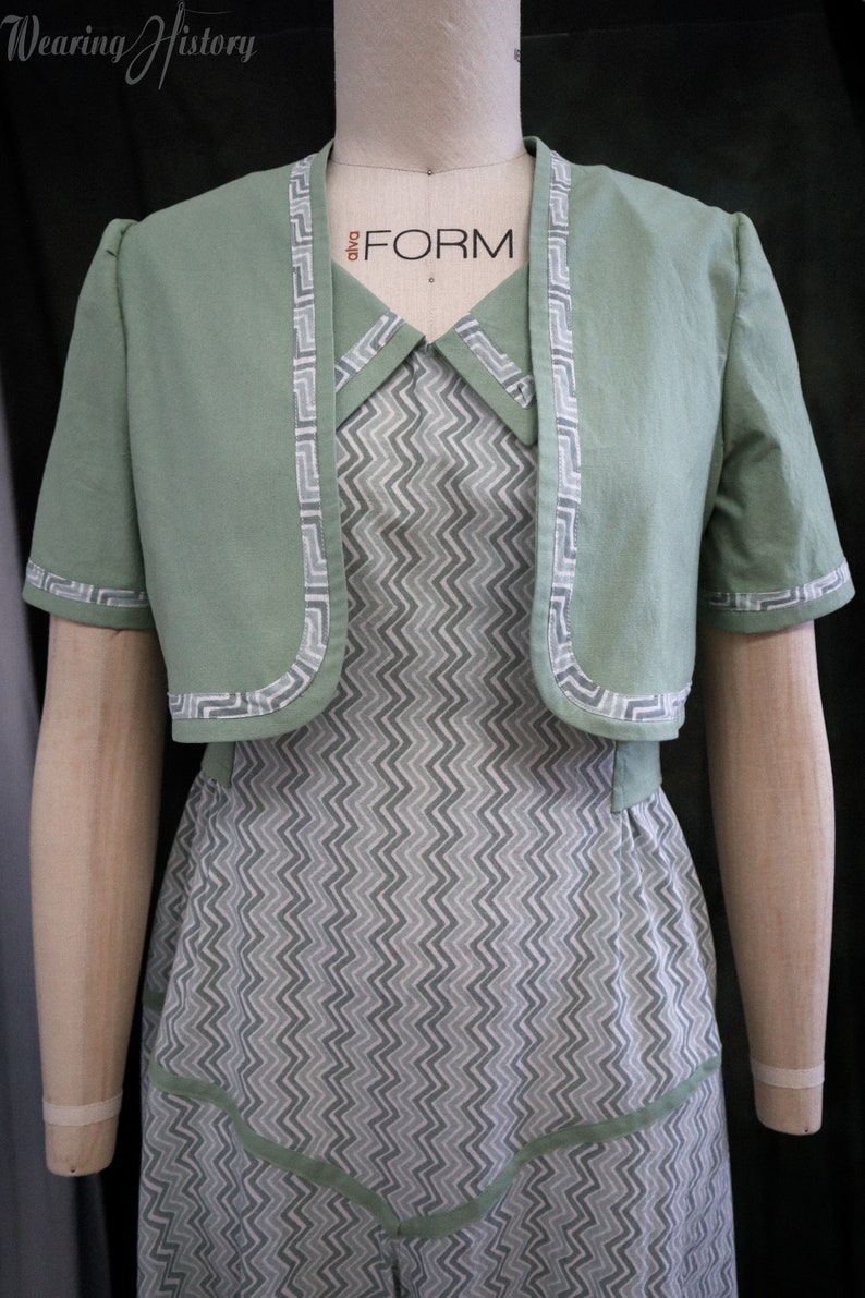 May include: A green cropped jacket with a white zig-zag trim over a white and gray chevron patterned dress. The dress has a white collar and a green trim around the neckline. The dress has a fitted bodice and a full skirt.
