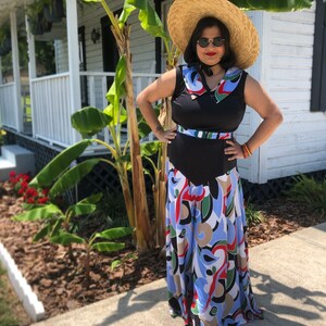 May include: A woman wearing a black sleeveless dress with a white and blue patterned skirt. She is wearing a wide-brimmed straw hat and sunglasses. The dress has a black belt and a white collar.
