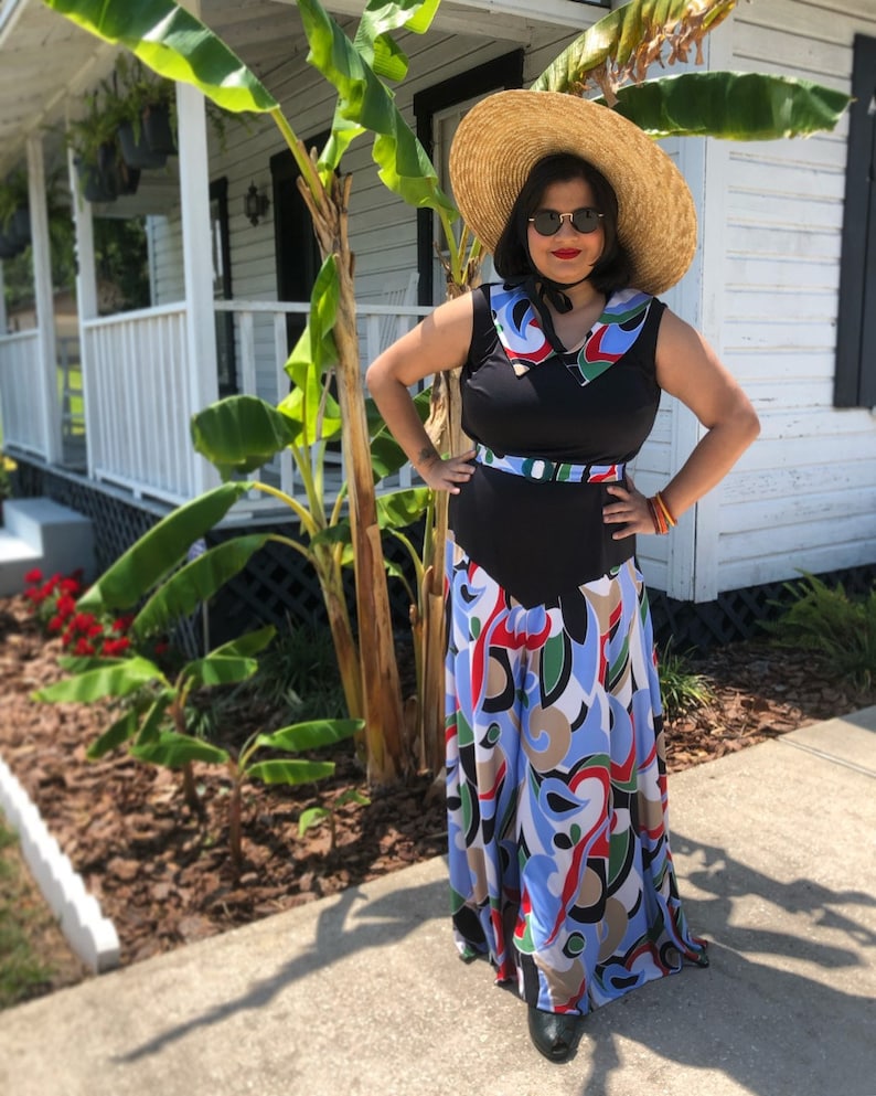May include: A woman wearing a black sleeveless dress with a white and blue patterned skirt. She is wearing a wide-brimmed straw hat and sunglasses. The dress has a black belt and a white collar.