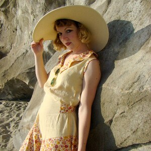 May include: A woman wearing a wide-brimmed straw hat, a yellow sleeveless dress with a floral print skirt, and a large bangle bracelet. The dress has a contrasting yellow bodice and a floral print skirt. The woman is standing next to a large rock.