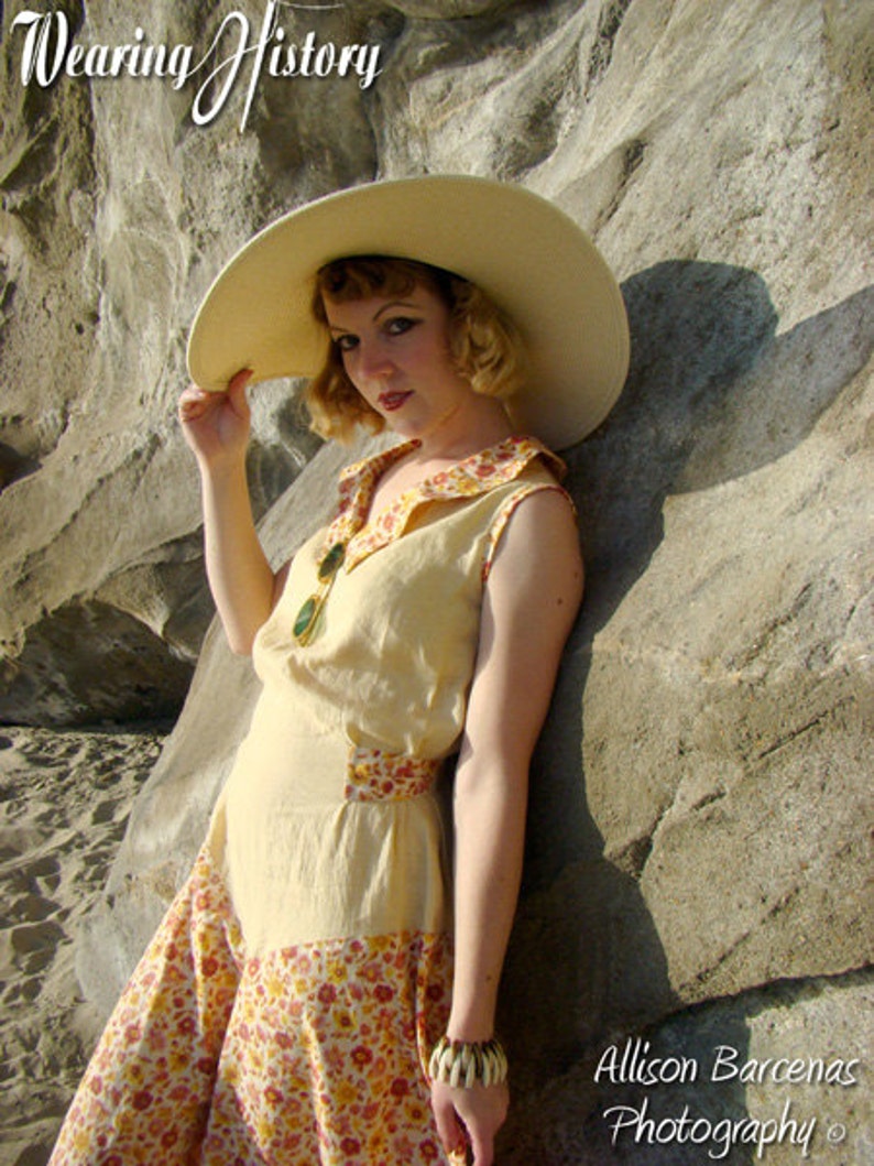 May include: A woman wearing a wide-brimmed straw hat, a yellow sleeveless dress with a floral print skirt, and a large bangle bracelet. The dress has a contrasting yellow bodice and a floral print skirt. The woman is standing next to a large rock.