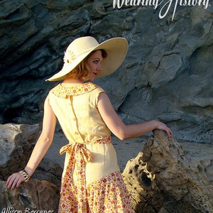 May include: A woman wearing a yellow linen dress with a floral print skirt and a wide-brimmed straw hat. The dress has a button-down back and a sash tied at the waist. The woman is standing on a rocky beach.