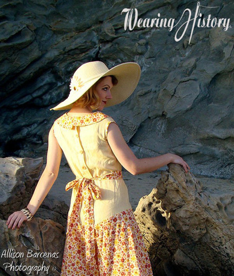 May include: A woman wearing a yellow linen dress with a floral print skirt and a wide-brimmed straw hat. The dress has a button-down back and a sash tied at the waist. The woman is standing on a rocky beach.