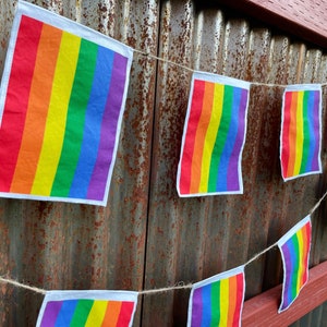 May include: A rainbow flag banner strung across a rusty metal wall. The banner is made of fabric and has six stripes of red, orange, yellow, green, blue, and purple.