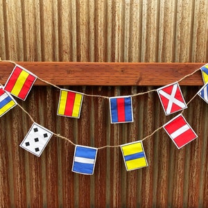 Puede incluir: Una hilera de banderas de señales náuticas colgando sobre un fondo de metal oxidado. Las banderas son rojas, amarillas, azules y blancas con varios símbolos y letras.