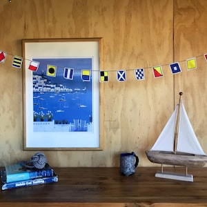 May include: A framed print of a harbor scene with sailboats hangs on a wood panel wall. A string of nautical flags hangs above the print. A small wooden sailboat model sits on a wooden table with a blue ceramic mug and two books.