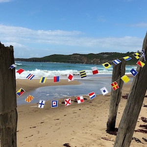 May include: A string of colorful nautical signal flags hangs between weathered wooden posts on a sandy beach. The flags, in various shapes and colors, are strung together against a backdrop of ocean waves and a blue sky.