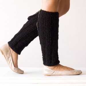 May include: Black knitted leg warmers with a textured pattern. The leg warmers are worn with beige ballet flats. The background is a plain white wall and floor. The leg warmers appear to be handmade.