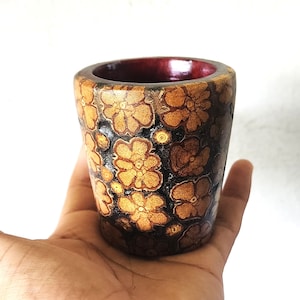 Op de afbeelding: Een klein, bruin keramiek kopje met een bloemenpatroon. Het kopje heeft een donkerbruine rand en is versierd met een herhalend patroon van gele bloemen op een zwarte achtergrond.