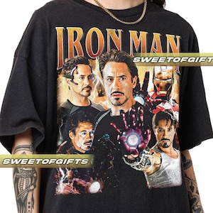 Könnte beinhalten: Schwarzes T-Shirt mit goldenem "IRON MAN"-Schriftzug und einer Collage von Iron Man-Bildern. Das Design zeigt mehrere Porträts der Figur, wobei das Gesicht und die Hand des Superhelden mit einer leuchtenden Energiequelle hervorgehoben werden.