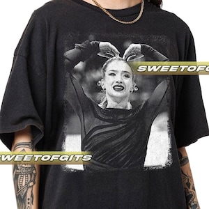 Puede incluir: Camiseta negra con una imagen en blanco y negro de una mujer con los brazos levantados, las manos formando un corazón. El texto "SWEETOFGITS" está impreso en la camiseta.