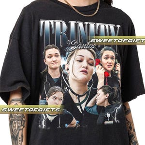 Könnte beinhalten: Schwarzes T-Shirt mit einer Grafik einer Frau mit Stethoskop und dem Text "TRINITY Santos" in silberner, stilisierter Schrift. Das Design enthält mehrere Bilder der Frau. Das T-Shirt hat kurze Ärmel.