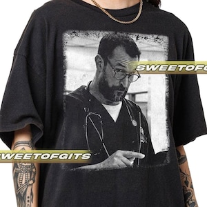 May include: Black t-shirt with a black and white image of a man in glasses and a medical uniform. The image is framed in a white border. The text "SWEETOFGITS" is printed across the shirt.