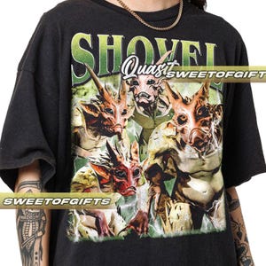 Puede incluir: Camiseta negra con un gráfico de cuatro criaturas fantásticas con piel roja, marrón y beige, y orejas grandes. La palabra "SHOVEL" está impresa en verde y blanco encima de las criaturas, con "Quasi" en el centro.