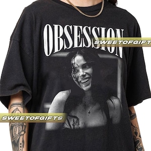 May include: Dark gray t-shirt with the word "OBSESSION" in white block letters above a black and white image of a smiling person. The image has blood-like marks on the face.