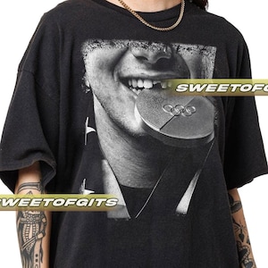 May include: Black t-shirt featuring a black and white graphic of a person biting a silver Olympic medal. The image is a close-up of the person's face. The text "SWEETOFGITS" is printed on the shirt.