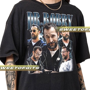 以下が含まれることがあります： ひげと眼鏡をかけた男性の画像をコラージュしたダークグレーのTシャツ。銀色の様式化されたフォントで「DR. BOBBY」の文字がプリントされています。ヴィンテージ感のあるTシャツで、「SWEETOFGITS」の文字がプリントされています。