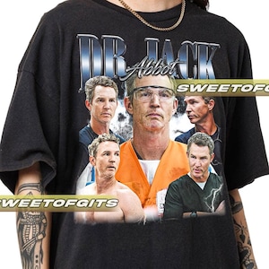 May include: Black t-shirt featuring a graphic of a man with the text "DR. JACK Abbot" in a silver and blue font. The graphic includes multiple images of the man in various settings. The shirt has a crew neck and short sleeves.