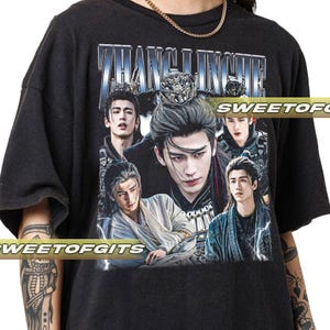 May include: Dark gray t-shirt featuring a collage of Asian men with long hair and ornate headwear. The shirt has the text "ZHANG LINGHE" in a silver and blue font. The brand name "SWEETOFGITS" is printed on the shirt.