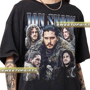 May include: Black t-shirt featuring a graphic of Jon Snow from Game of Thrones. The design includes multiple images of the character with the name "JON SNOW" in a silver and blue font. The text "SWEETOFGIFTS" is also visible.