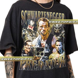 May include: Black t-shirt with a graphic print of Arnold Schwarzenegger and the Predator from the movie Predator. The text "Schwarzenegger" and "Predator" are printed on the shirt.