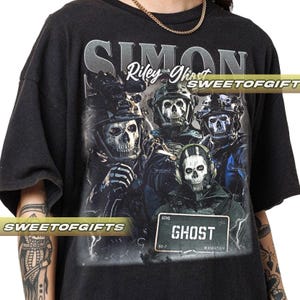 Könnte beinhalten: Schwarzes T-Shirt mit einer Grafik von vier soldatischen Schädelgesichtern in taktischer Ausrüstung. Das Design enthält den Text "SIMON Riley Ghost" und ein Straßenschild mit der Aufschrift "GHOST" und "Manhattan."
