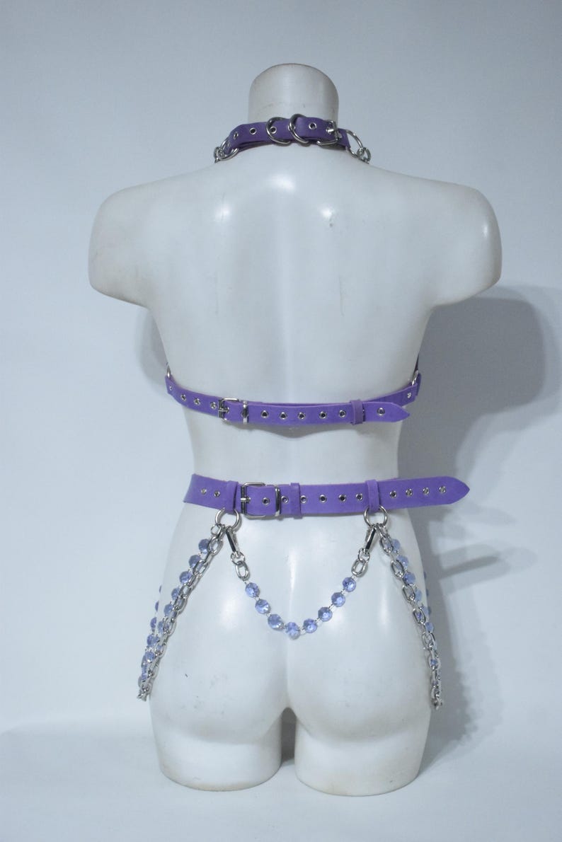 May include: A lavender purple harness set featuring a choker, a chest strap, and a waist belt with silver-toned metal buckles and eyelets. The waist belt is adorned with dangling silver chains and light purple faceted crystal beads.