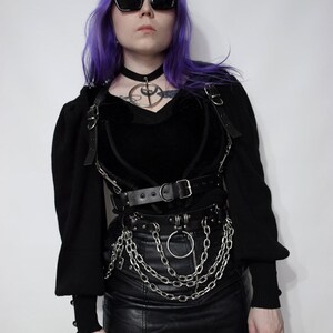 May include: A woman with purple hair wearing a black choker necklace, a black velvet heart-shaped top, a black leather skirt, and a silver chain belt with a large silver ring.