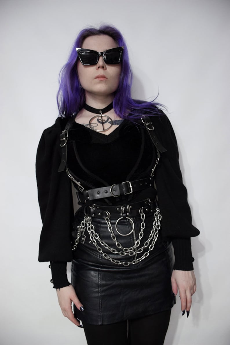 May include: A woman with purple hair wearing a black choker necklace, a black velvet heart-shaped top, a black leather skirt, and a silver chain belt with a large silver ring.