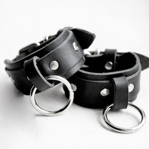 Puede incluir: Dos puños de muñeca de cuero negro con anillos y remaches de metal plateado.