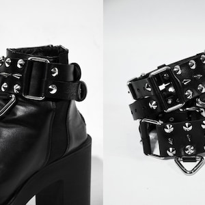 May include: Two black leather belts with silver buckles and metal studs. The belts are layered on top of each other and have a triangular metal loop on the end.
