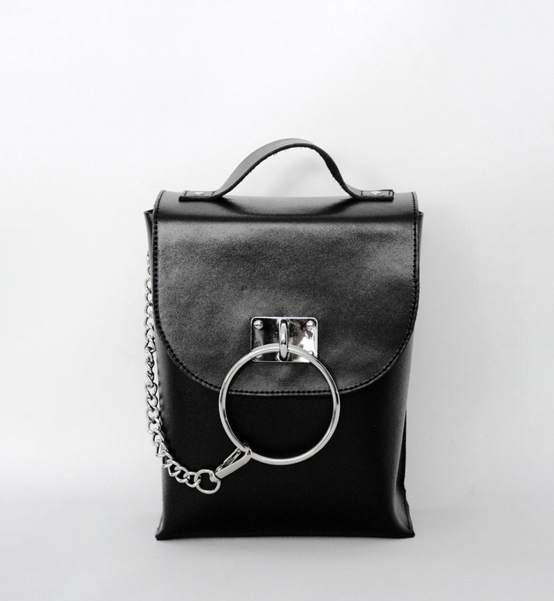 Puede incluir: Una mochila de cuero negro con un detalle de anillo y cadena plateados. La mochila tiene un cierre de solapa y un asa superior.