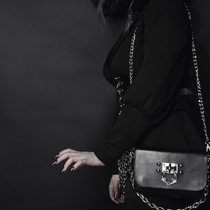 May include: A black purse with a silver chain strap and a silver clasp. The clasp has a decorative design with a skull and wings.