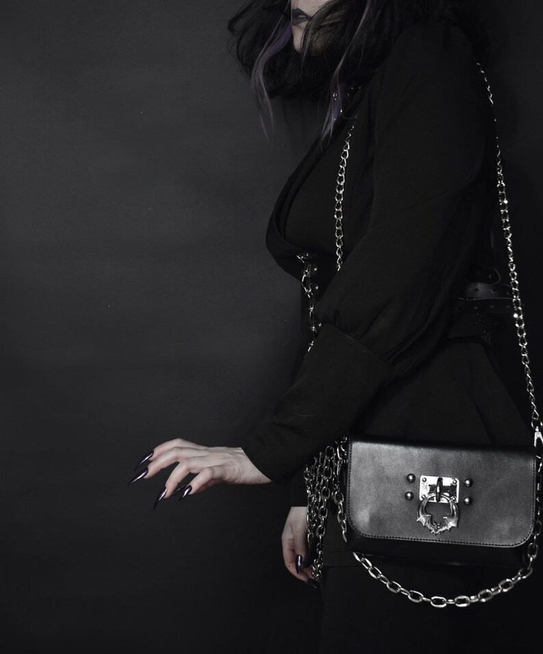 May include: A black purse with a silver chain strap and a silver clasp. The clasp has a decorative design with a skull and wings.