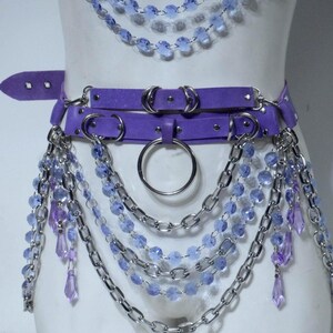 May include: A purple harness set featuring a top piece and a waist belt adorned with silver-toned metal chains, circular rings, and dangling light blue and purple faceted crystal-like beads that drape across the torso and hips.