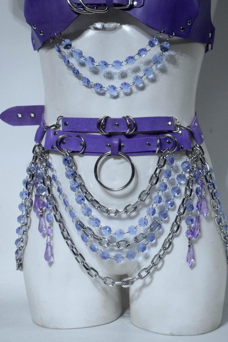 May include: A purple harness set featuring a top piece and a waist belt adorned with silver-toned metal chains, circular rings, and dangling light blue and purple faceted crystal-like beads that drape across the torso and hips.