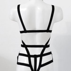 May include: Black body harness with a choker neckline. The harness has multiple straps forming a geometric pattern across the torso and back. The garment is displayed on a white mannequin.