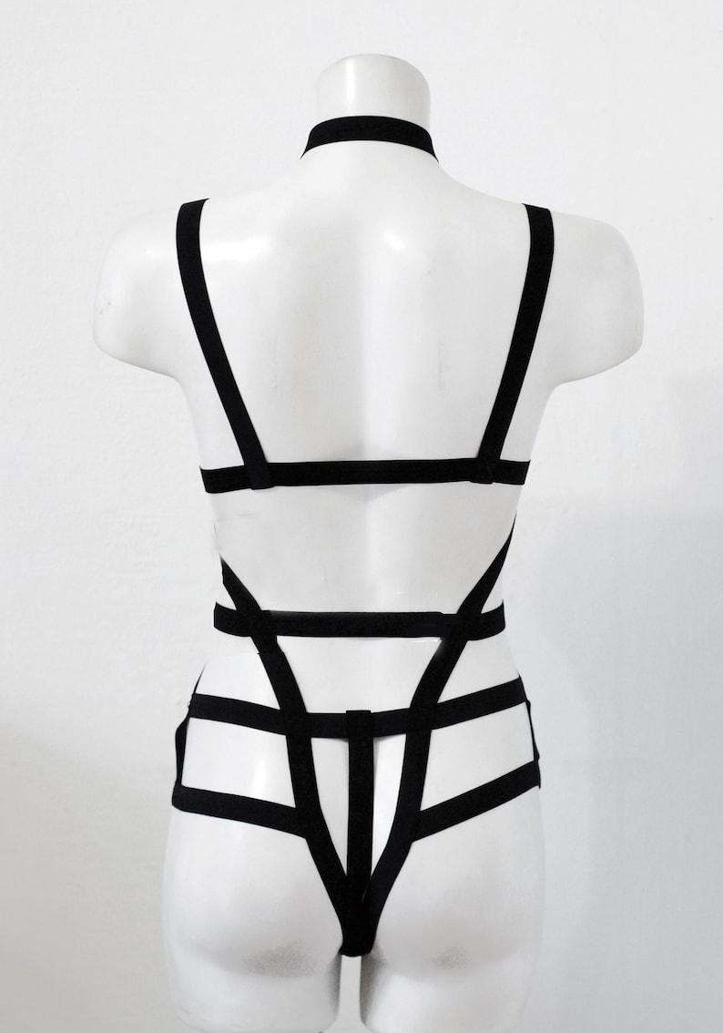 May include: Black body harness with a choker neckline. The harness has multiple straps forming a geometric pattern across the torso and back. The garment is displayed on a white mannequin.