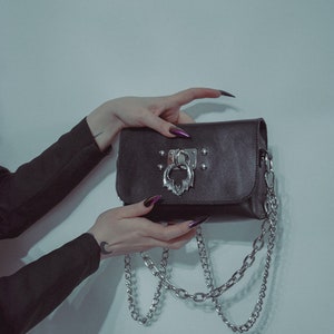 May include: A black leather purse with a silver chain strap and a silver clasp with a bat design. The purse has silver studs around the edges.
