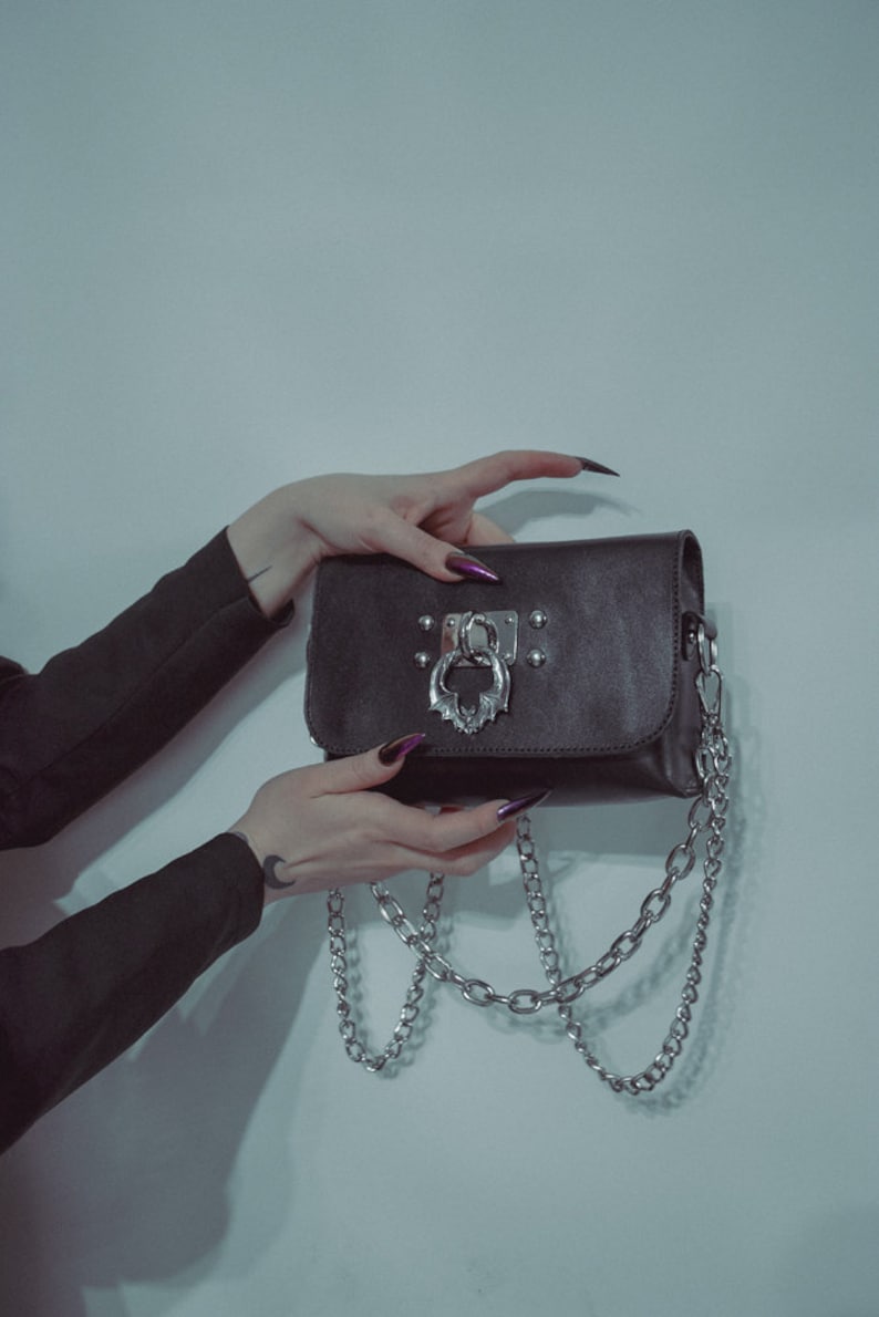 May include: A black leather purse with a silver chain strap and a silver clasp with a bat design. The purse has silver studs around the edges.