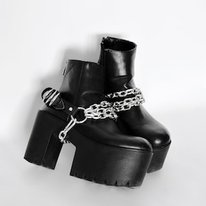 Może przedstawiać: Czarne buty platformowe z srebrnymi akcentami łańcucha. Buty mają gruby obcas i grubą platformę.