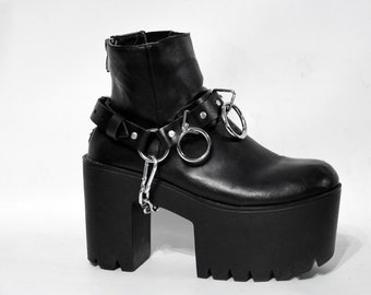 Könnte beinhalten: Schwarze Plateau-Stiefeletten aus Leder mit einem dicken Absatz und silbernen Kettendetails. Die Stiefeletten haben einen Riemen mit silbernen Ringen um den Knöchel.