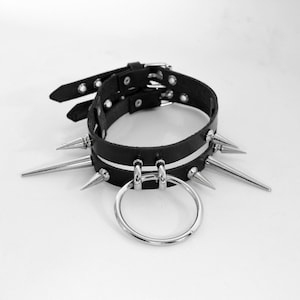 May include: A black leather choker with silver spikes and a silver ring. The choker has a white stripe running around it.
