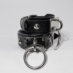 Könnte beinhalten: Schwarze Lederhandgelenkbänder mit silbernen Metallakzenten, darunter ein großer Ring und ein kleiner D-Ring. Die Manschetten sind mit silbernen pyramidenförmigen Nieten besetzt.
