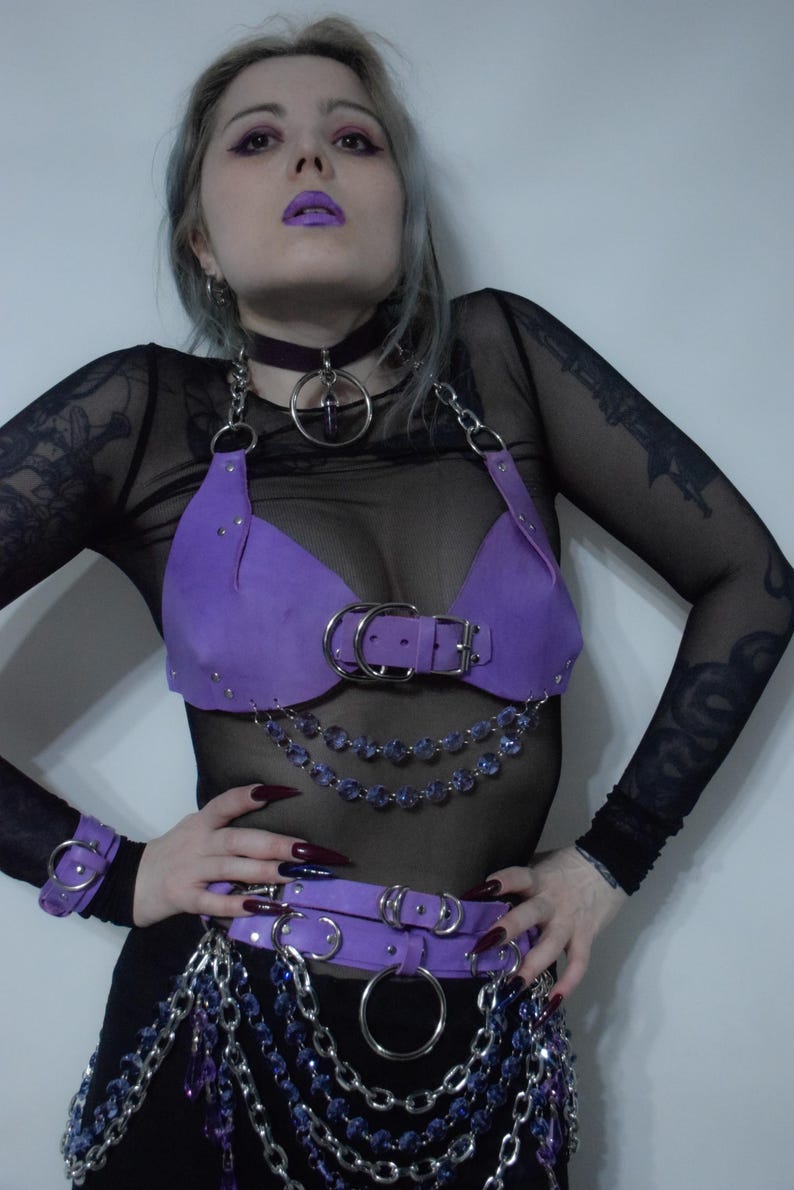 May include: A purple leather-like chest harness featuring silver-toned metal buckles, O-rings, and studs. The harness includes draped chains with purple faceted beads, a matching waist belt with hanging chain fringe, and a wrist cuff accessory.