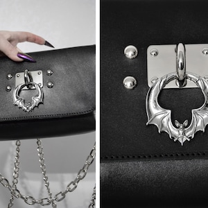 May include: Black leather crossbody bag with a silver bat-shaped clasp and a silver chain strap.