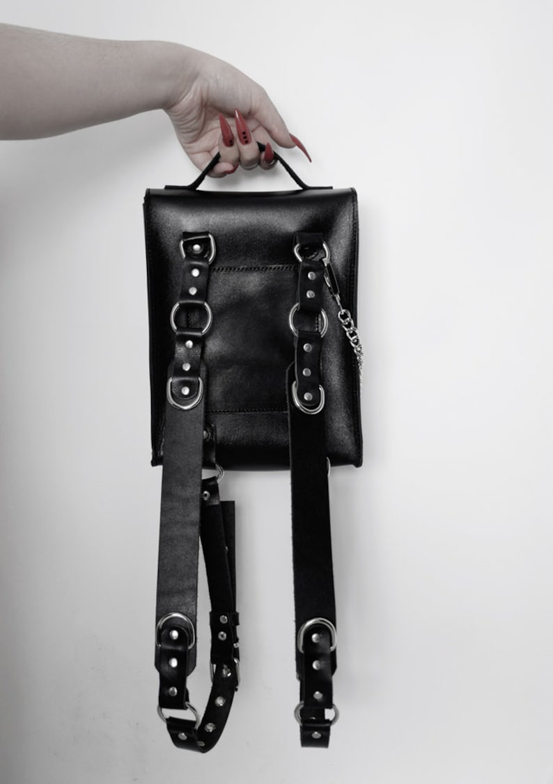Puede incluir: Mochila de cuero negro con herrajes plateados y correas ajustables. La mochila tiene m&uacute;ltiples hebillas y un detalle de cadena.