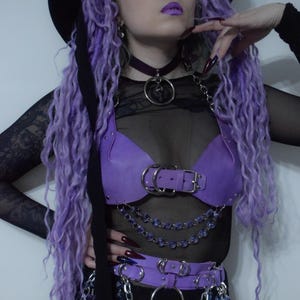 May include: A purple leather-like chest harness and matching waist belt featuring silver-toned metal buckles, O-rings, and layered chains with purple faceted beads. The set is worn over a black sheer long-sleeved top and a black choker with a pendant.