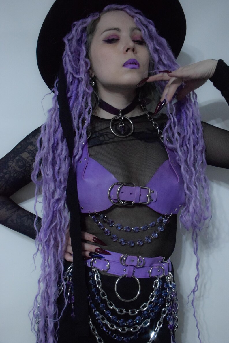 May include: A purple leather-like chest harness and matching waist belt featuring silver-toned metal buckles, O-rings, and layered chains with purple faceted beads. The set is worn over a black sheer long-sleeved top and a black choker with a pendant.