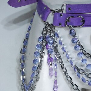 May include: A purple suede belt featuring silver-toned metal hardware, including circular rings and a buckle. The belt is adorned with multiple draped silver chains and strands of faceted light blue and purple crystal-like beads and pendants.